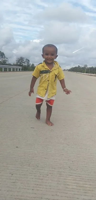 Child in yellow shirt walking on a gray road under a cloudy sky.