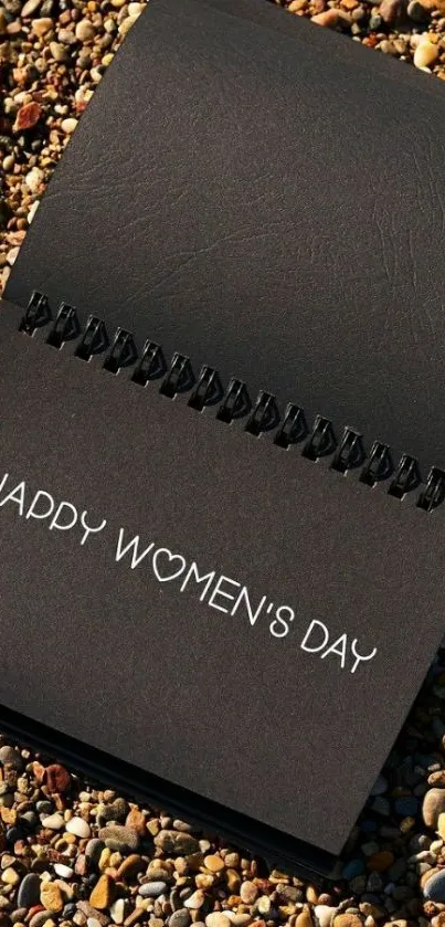 Elegant black notebook with Happy Women's Day message on pebbles.