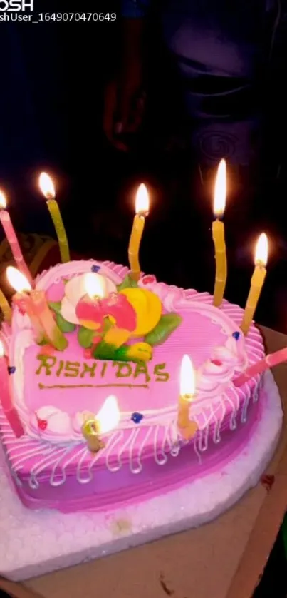 Heart-shaped cake with pink icing and lit candles on top.