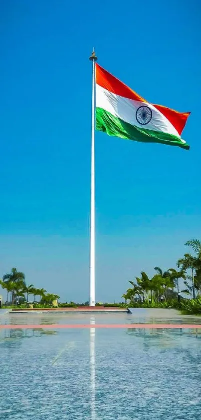 Indian flag under a clear blue sky, tall flagpole reflected in water.