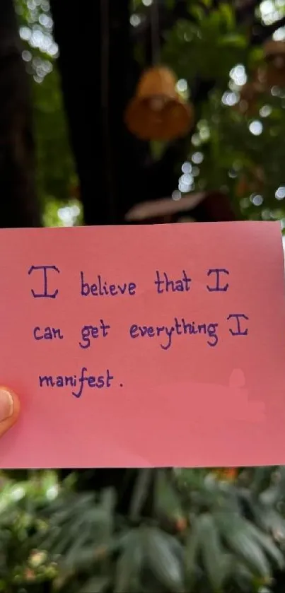 A motivational handwritten note on pink paper set against a lush green background.