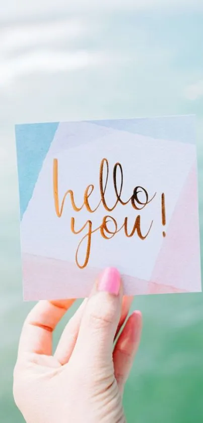 Hand holding a colorful greeting card with 'hello you!' by the ocean.