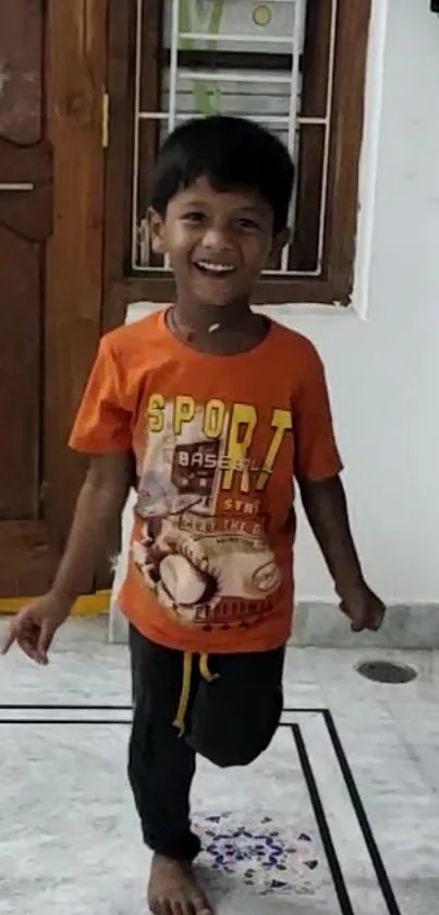 Smiling child in orange shirt standing joyfully indoors.