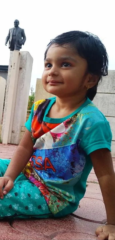 A joyful child in colorful clothes sitting on a paved ground outdoors.