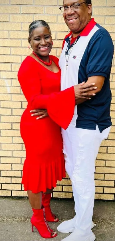 Couple embracing happily in red outfit against brick wall.