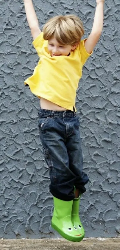 Child in yellow shirt jumping joyfully against grey wall.