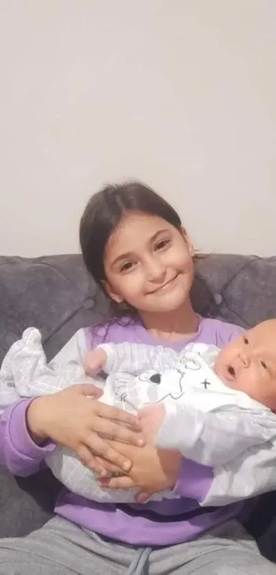 A smiling child holds a baby on a gray couch, showcasing a loving sibling bond.