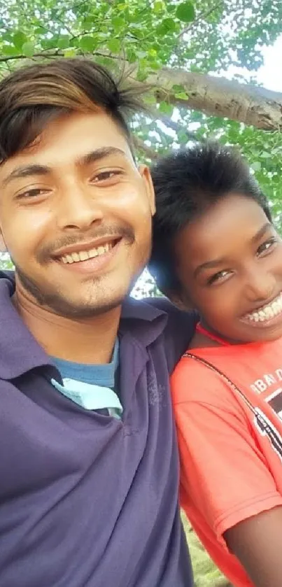 Two friends smiling joyfully under green trees.