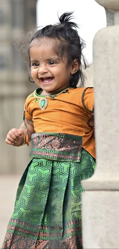 Smiling toddler in festive attire peeking around a pillar.