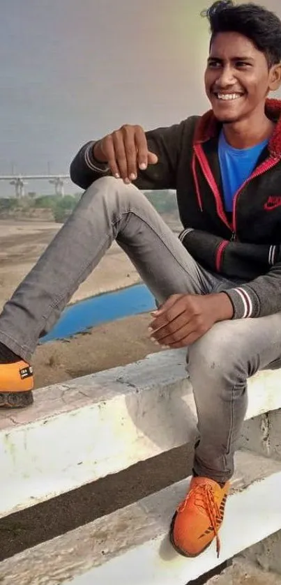 Smiling young man sitting outdoors in casual attire with vibrant city backdrop.