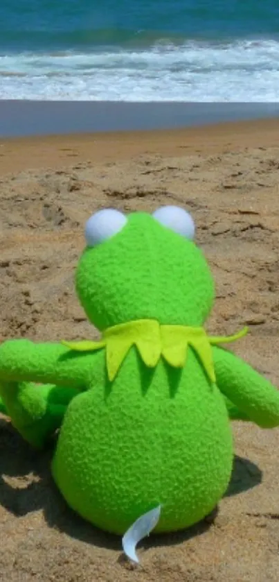 Green frog toy sitting on the sandy beach with ocean waves in the background.