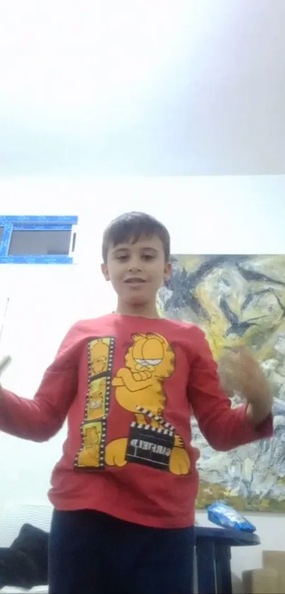 A young boy wearing a red cartoon shirt stands indoors with colorful artwork behind him.