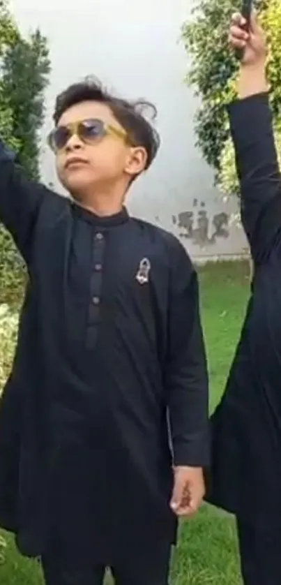 Two kids in black traditional outfits raising hands in a green garden.