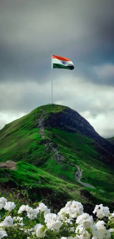 A colorful flag waves on a green mountain under a dramatic sky with white flowers below.