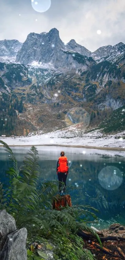 Person standing by a tranquil mountain lake with snowy peaks and lush greenery.