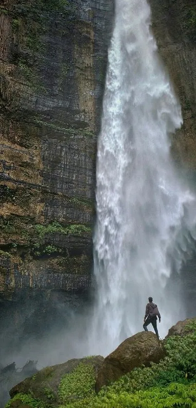 Man standing by a majestic waterfall amidst lush greenery and rocky cliffs.
