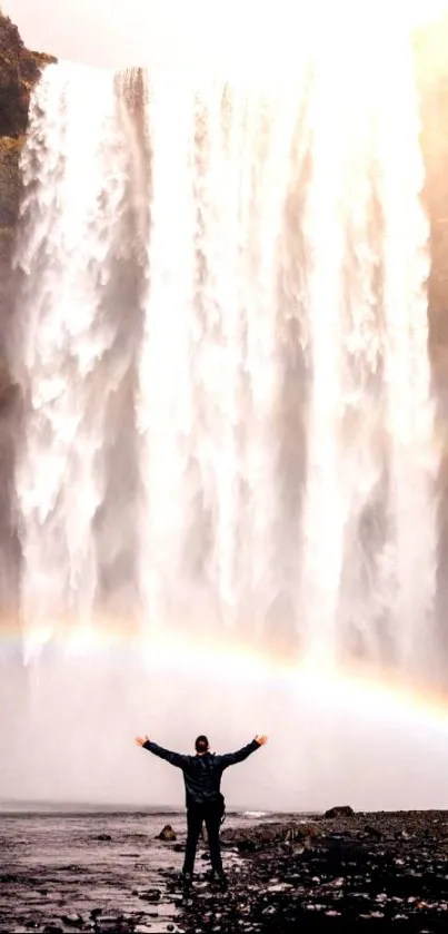 Man stands before a majestic waterfall with a vivid rainbow overhead.