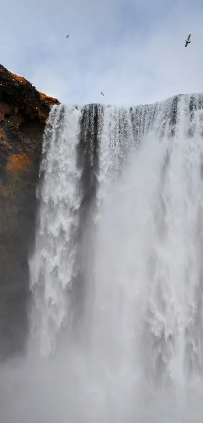 Majestic waterfall cascading down cliffs with a lone figure nearby, under a blue sky.