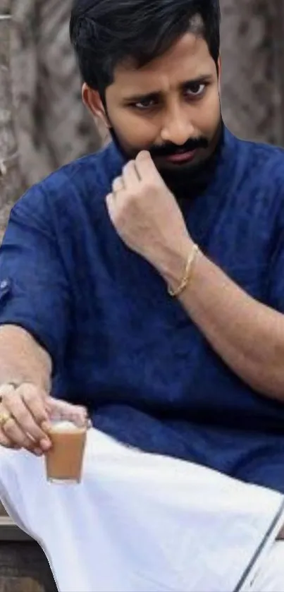 Man in traditional blue attire holding a drink, set in a rustic backdrop.