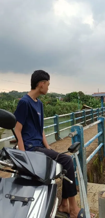 Man on motorcycle by a bridge under cloudy skies.