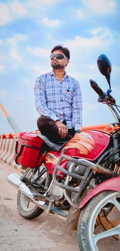 Man sitting on red motorcycle by a bridge under blue sky.