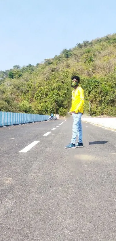 Man in yellow jacket walking along scenic road with green hill background.