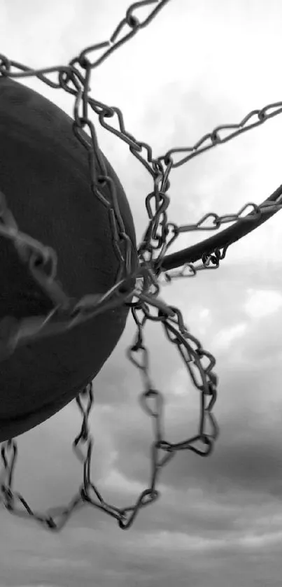 Monochrome image of basketball hoop with ball against cloudy sky.