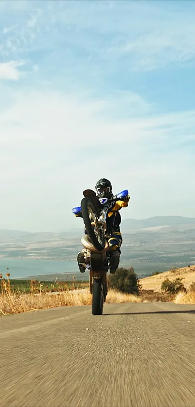 Motorcyclist performing a wheelie on a scenic road with clear blue sky.