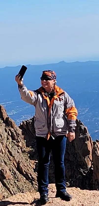 Person holding phone on rocky mountain with blue sky.