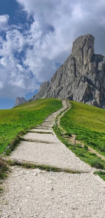 Path leading up a green meadow towards a towering rocky mountain under a blue sky.