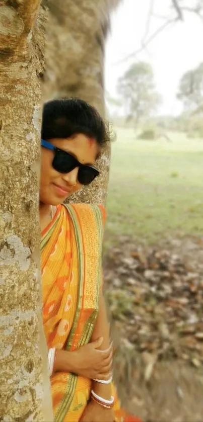 Woman in orange sari by a tree, wearing sunglasses.