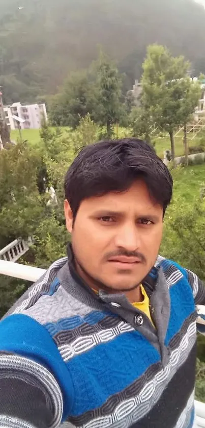 Man in a sweater taking a selfie with greenery and buildings in the background.