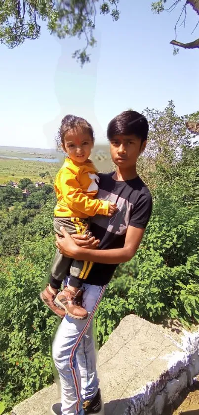 Boy holding child in lush green setting with clear skies.