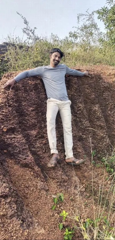 Man posing on a brown cliff with greenery in the background under a clear sky.