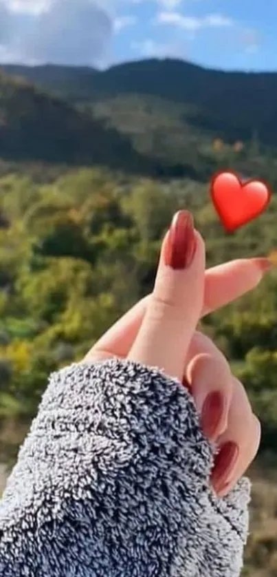 Hand gesture with heart over scenic green landscape.