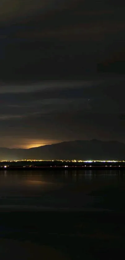 Nighttime view of mountains with city lights and reflection on a calm lake.