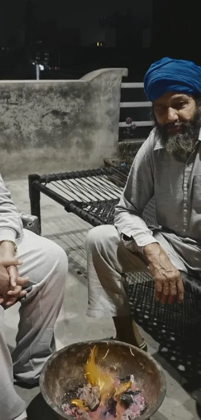 Two men in traditional attire on a rooftop, sitting by a fire at night.