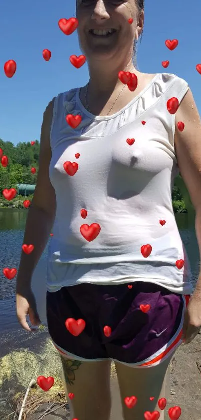 Smiling person by a lakeside with floating red hearts under a clear blue sky.