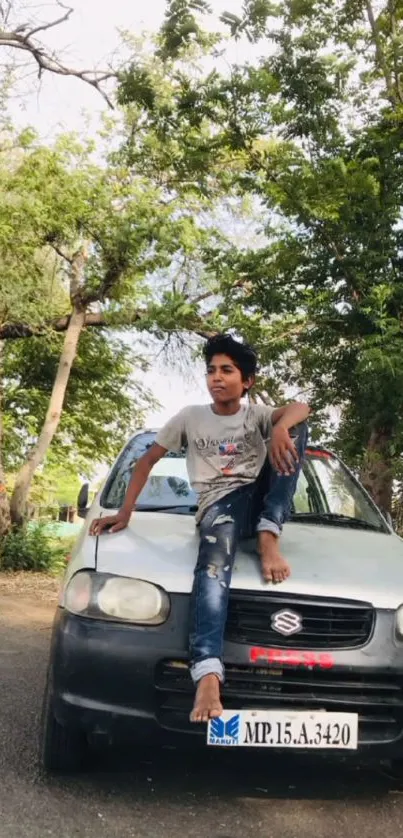 Young man sits on car beneath green trees.