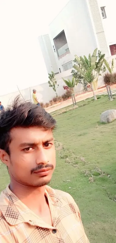 Young man takes selfie in green garden with modern building backdrop.