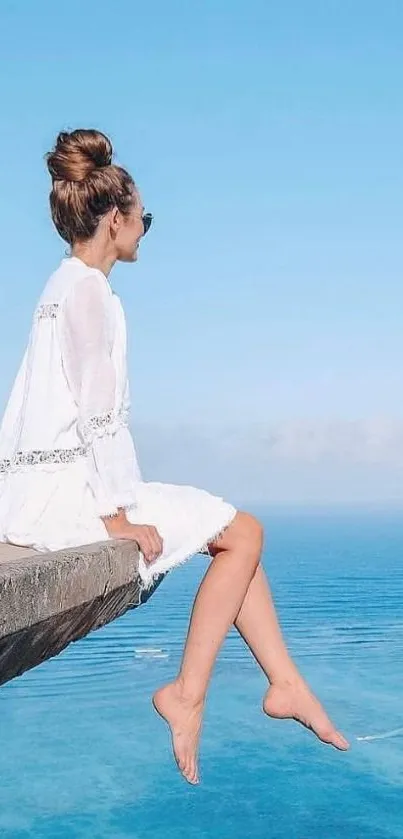 Woman in white dress sitting on a cliff overlooking the ocean.