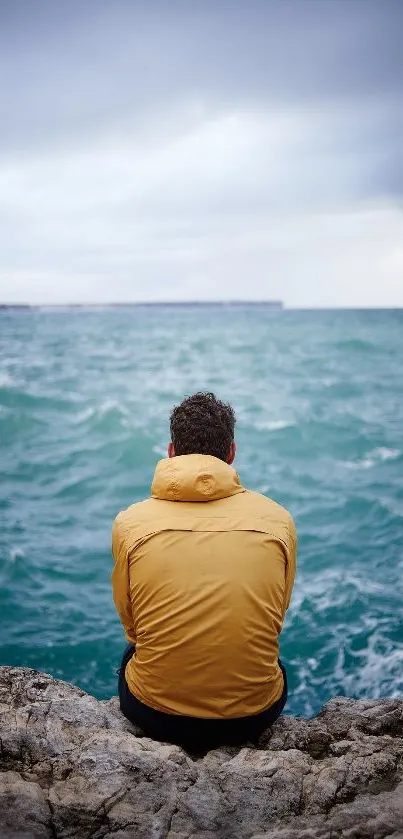 Person in yellow jacket sitting by the ocean, gazing at the sea and sky.