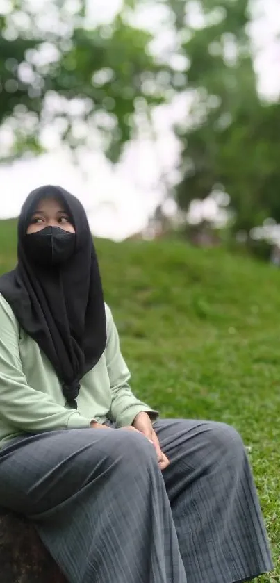 Woman in hijab sitting on a stone in a lush green park wearing a mask.