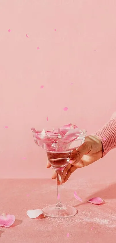 Hand holding a glass with pink rose petals on a soft pink background.