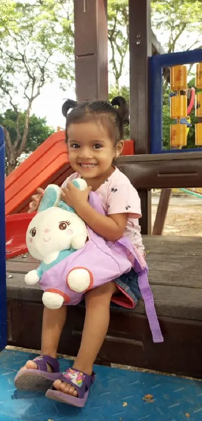 Child sitting on a playground slide with a plush toy, smiling joyfully.