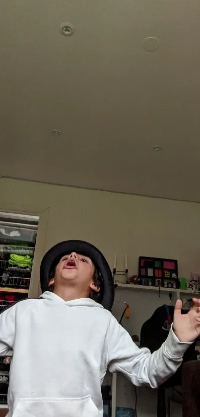 Child in white hoodie playfully posing indoors with a hat on.