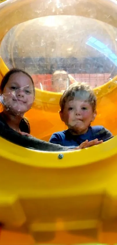 Two children smiling inside a yellow play capsule.