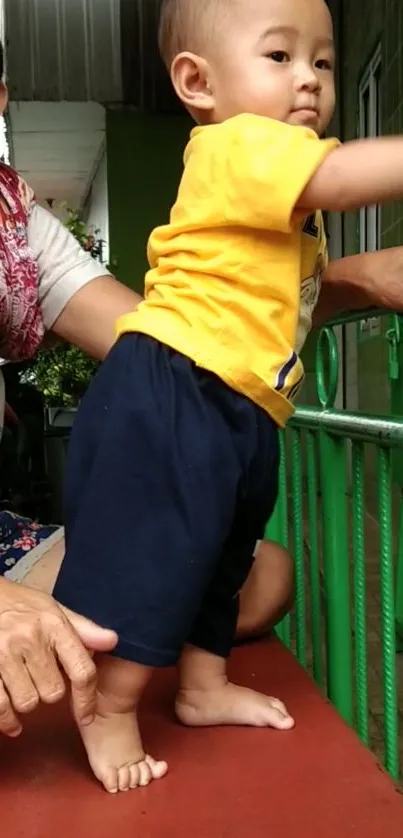 Young toddler standing next to a green railing, wearing a bright yellow shirt.