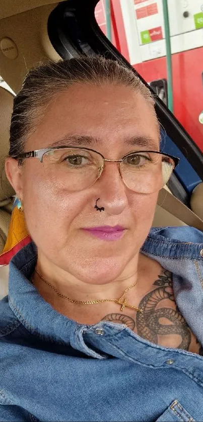 Woman sitting in car with denim shirt and glasses, showing tattoos and a calm expression.