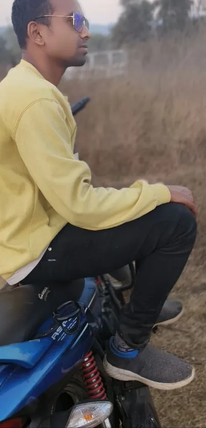 Young man in yellow shirt sitting on a blue motorcycle in nature.